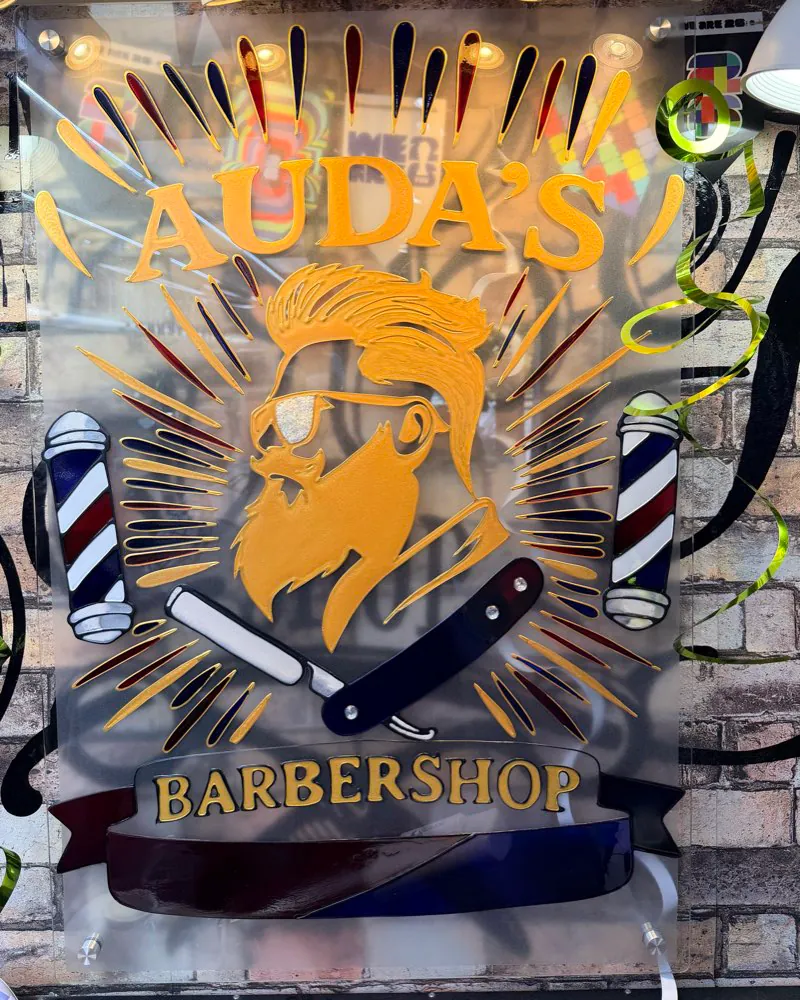 Reverse glass painted barbershop sign with a gilded bearded barber portrait, twin barber poles, crossed straight razors, and a ribbon banner reading Auda's Barbershop